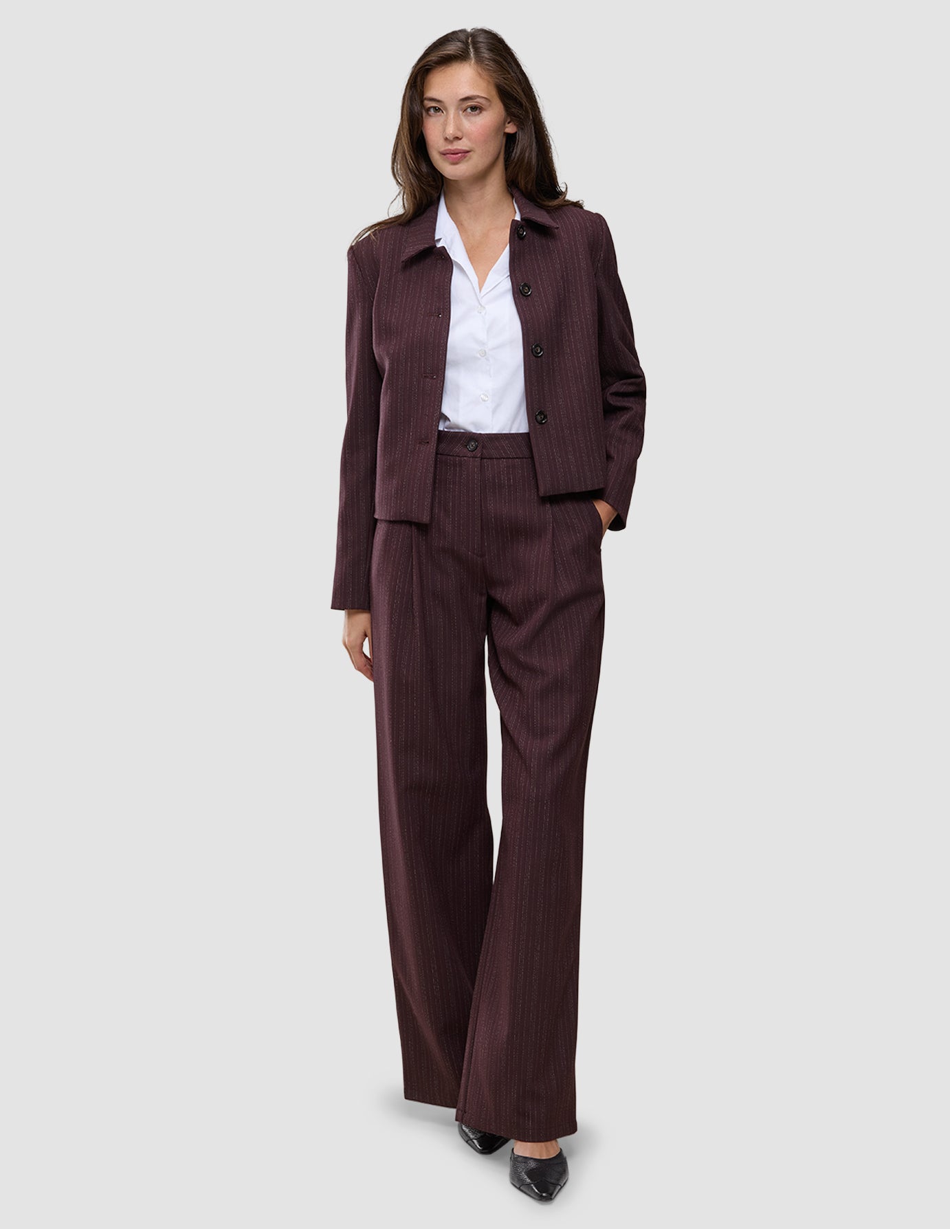 En kvinde iført et buksesæt af modellen Women Serene Pants Wide Berry Pinstripe over en hvid skjorte med krave.